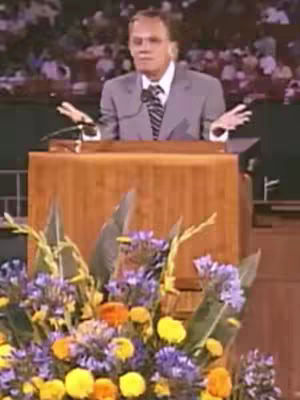 An older man speaking at a podium, with his hands open as if sharing a message. Behind him, there's an audience blurred out of focus. In front of the podium are colorful flowers.