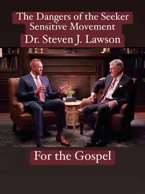 Two men in suits are seated in a warmly lit room, likely a study or library. They are facing each other, engaged in conversation, with text overlays indicating the video's topic. The setting includes a fireplace, bookshelves, and armchairs, suggesting a formal interview or discussion.