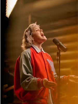 A female singer with short blonde hair, dressed in a red and gray baseball jacket, is singing with passion into a microphone. The lighting is warm and the background is an out-of-focus studio.