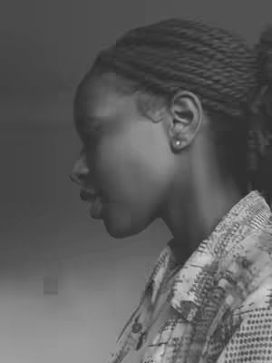 A grayscale image showing the side profile of a woman with braided hair pulled back into a bun. She's wearing a patterned shirt with the collar slightly visible. The background is a gradient of gray, with good lighting on the woman's face. The focus is on her features, especially the nose, lips, and ear.