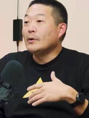 A man in a black shirt, with a black microphone in front of him, is speaking. The man is Asian with short black hair, a small mustache, and is wearing a black watch on his left wrist. Behind the man, is a dark square, which is likely an electrical box.