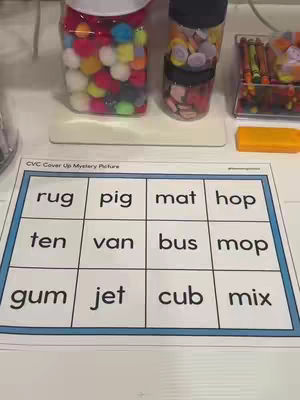 The image shows a classroom setting with educational materials. On a white surface, there are jars filled with colorful pom-poms, bottle caps, and crayons. In the foreground, a worksheet with CVC words and a grid is visible.