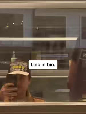 A person is reflected in a window, holding a phone up to take a picture. The image is taken at night, with the background out of focus, creating a sense of mystery. A sticker with the text 'Link in bio.' is placed below the window.