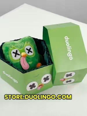 A plush green owl, modeled after the Duolingo mascot, is displayed inside an open green box that resembles a coffin. The owl has white 'x' eyes, a yellow beak, and a pink tongue sticking out. The box has white 'x' eyes as well, with pink tongues to mimic the owl and text 'store.duolingo.com' is written on the bottom with yellow letters. The background appears to be a white surface.