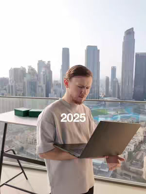 A man standing on a balcony with a laptop open in front of him, with a cityscape skyline in the background under a bright sky, and the year 2025 overlaid as text.