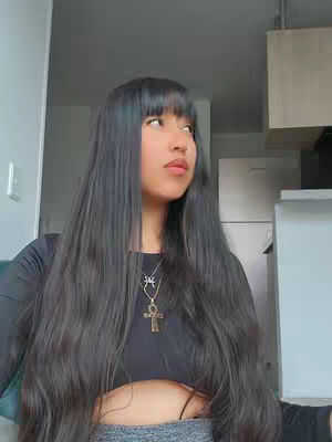 A young person with long dark hair, bangs, and a black top looks slightly off-camera. They are wearing a necklace with a cross pendant.