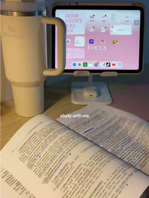The image shows a study setup with an open book in the foreground, a white insulated tumbler to the left, and a tablet on a stand displaying a pink background with app icons behind it. The words "study with me" are overlaid on the image.