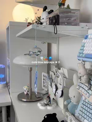 A well-lit, aesthetically pleasing desk setup with a white shelf holding various items, including a keyboard, decorations, and accessories. In the foreground, a decorative hanging piece and several figurines add visual interest. Soft lighting and a focus on pastel colors enhance the cozy and inviting atmosphere.