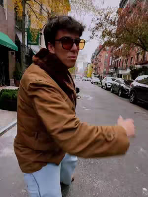 A man wearing sunglasses and a brown jacket walks on a city street with buildings in the background. He gestures to his left.