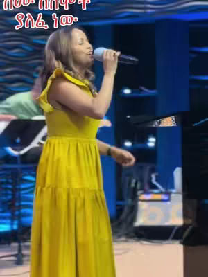 A woman in a yellow dress sings into a microphone on a stage with blue and black decor.