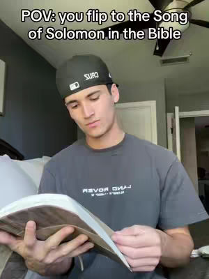 A young man in a grey t-shirt and black baseball cap is seated indoors, holding open a Bible. The text on the image reads, 'POV: you flip to the Song of Solomon in the Bible'. The room has a ceiling fan and white door frames.
