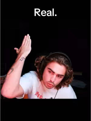 The cover image features a young man in a white t-shirt, holding up his left hand with the palm facing outward. Above him, in bold white text, is the word "Real." The man is wearing headphones and appears to be indoors, likely in front of a blurred background. The lighting is even, and the color palette is muted. A tattoo is visible on his left forearm. The overall composition is centered, with the upper half of the frame dominated by a black void.