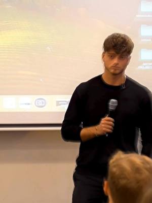 A man in a black long-sleeve shirt stands in front of a projection screen. He holds a microphone. There's an audience partially visible in the foreground.