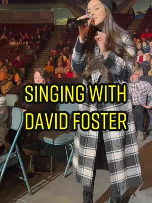 A woman in a black hat and checkered coat singing into a microphone in front of a crowd. The text overlay says, "SINGING WITH DAVID FOSTER".
