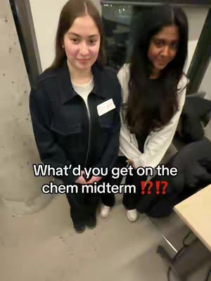 Two young women stand in a building, looking slightly down at the camera. The woman on the left is closer, wearing a black jacket over a white shirt. The woman on the right is wearing a white hoodie, and has long dark hair. Text overlayed on the image asks "What'd you get on the chem midterm !?!?"