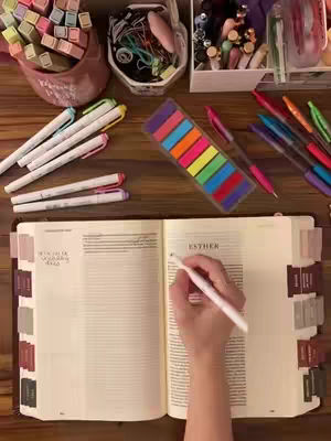 A top-down shot of a wooden table with various items related to bible study and journaling. The image is well-lit with natural light. The composition includes an open Bible, pens of different colors, sticky notes, and other stationery items. The overall look is organized and inviting.