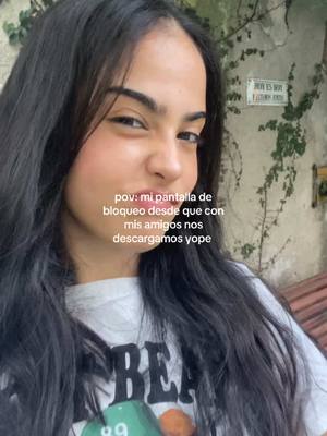 A selfie-style image features a young woman with long, dark hair centered in the frame. She is smiling slightly, looking towards the camera with a relaxed expression. She wears a white t-shirt with a graphic on it, part of which is visible. The background is somewhat blurry, with hints of foliage and a wall suggesting an outdoor setting. Overlayed text in the image reads, 'pov: mi pantalla de bloqueo desde que con mis amigos nos descargamos yope'.