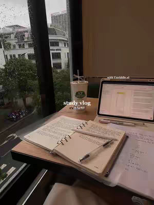 A well-lit image captures a student's study setup near a large window. A notebook and open book are on a wooden surface, with a laptop and a Starbucks cup, and the rainy view outside.