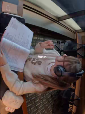 A woman wearing headphones studies a book while holding a pen in a recording studio. The image is well lit with neutral tones, emphasizing the subject's focus.