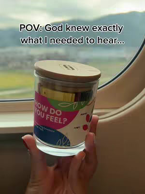A hand holding a glass jar with a wooden lid, containing various items, with a label that reads 'HOW DO YOU FEEL?'. The background shows a window with a blurred view of green landscape and a cloudy sky, likely from an airplane or train.