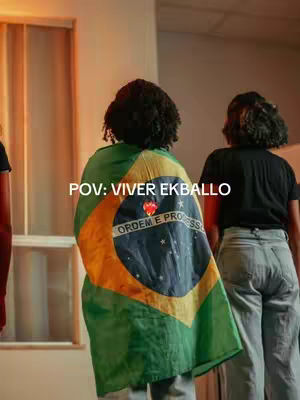 The image shows two persons from the back side, with one of them wearing a Brazilian flag, covering their back and part of the shoulders. The person wearing the flag has curly black hair. The image has a warm color palette, with the flag colors being the most prominent (green, and yellow). The text "POV: VIVER EKBALLO" is added on the cover.