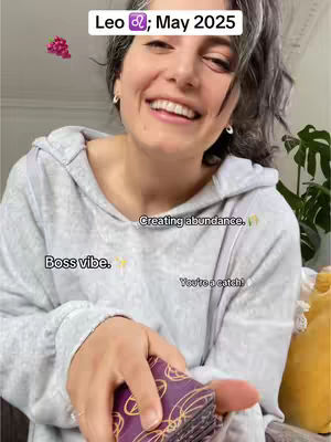 A woman with gray hair smiles, holding a tarot deck. Text overlays read 'Leo ♌; May 2025', 'Boss vibe,' 'Creating abundance,' and 'You're a catch!' A pink, flower-like graphic is also visible.