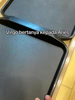 The cover image shows two black trays, possibly used for serving or carrying items, positioned diagonally in the frame. The trays have a textured surface and a black rim. A hand is visible, touching the tray on the bottom right. Overlayed text in white reads, 'Virgo bertanya kepada Aries.' The overall composition employs a simple, uncluttered design.