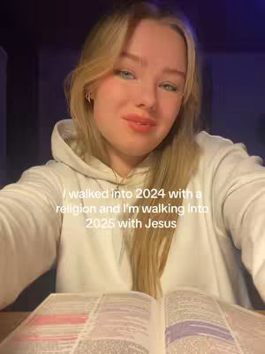 A young woman with blonde hair and a white hoodie is holding an open book in front of her. Text overlaying the image states, 'I walked into 2024 with a religion and I'm walking into 2025 with Jesus.' The background is dark, providing contrast to the woman's light features and clothing.