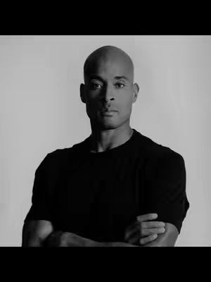 A black and white photo featuring a bald man in a black shirt with his arms crossed. The background is a plain, light color, making the man the central focus.