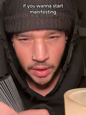 A close-up shot of a man wearing a black beanie and hoodie, looking intently at the camera. Text overlayed says, "if you wanna start manifesting". The background is dark, with a hint of a lit object on the right.