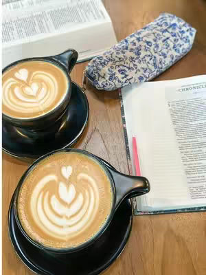 The cover image shows a top-down view of two coffee cups with latte art hearts, placed on saucers. Next to them are open books and a floral patterned mask. The setting appears to be a wooden table, with the lighting suggesting a well-lit indoor space.