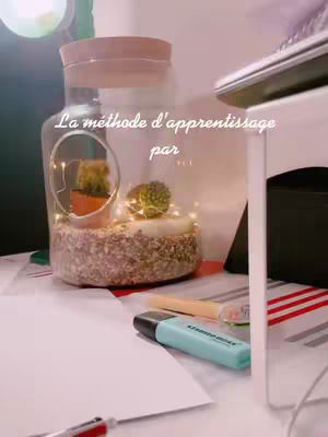 The image shows a close-up of a desk with a decorative glass jar containing succulents and fairy lights, along with a teal highlighter and a piece of paper. The composition is well-lit with a subtle focus on the study environment .