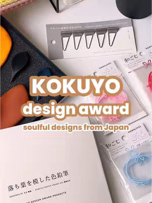 An overhead shot showcasing stationery products. The composition is well-lit, with a slight tilt.  At the bottom right, there are multiple stationery items in transparent packaging and products on a desk of a light color. In the center, overlaid text in a brown gradient announces "KOKUYO design award" . The overall tone is organized and appealing.