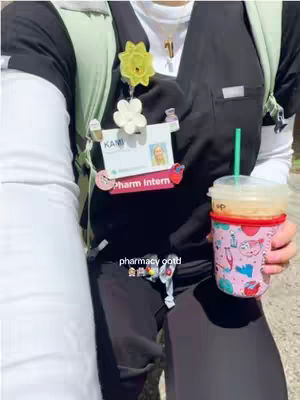 The image shows a close-up of a person, likely female, wearing black scrubs, with a light green backpack strap visible on the left. A name tag that says 'pharm intern' is attached to the scrubs. The person is holding a Starbucks iced coffee cup in their right hand. The cup has a pink and patterned design with drawings of objects. The text 'pharmacy gptd' and some emoji is in the bottom section of the image.