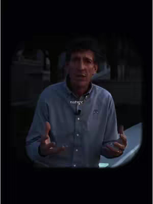A man in a light blue button-down shirt is positioned in the center of the frame, set against a dark background with blurred elements suggesting an outdoor setting. The man is looking directly at the viewer, and his hands are gesturing outward as if he is speaking. Overlayed at the bottom center of the shot is the word 'nature', implying a connection to the environment or the topic being presented. The image is framed with a dark oval vignette, which concentrates the viewer's focus on the man and his immediate surroundings.