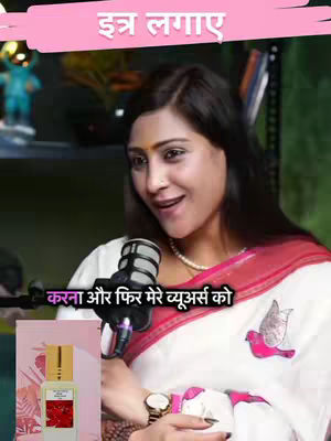 The image features a woman sitting in front of what appears to be a green screen. She is wearing a white saree with a red border and a pearl necklace, and she is also wearing a watch. There is a microphone in front of her, and a bottle of perfume is placed in the foreground. The background features a shelf with some items and other background objects. The overall composition is well-lit, and the focus is on the woman.
