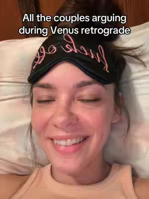 A selfie-style shot featuring a woman lying in bed, looking directly at the camera. She is wearing a black sleep mask with pink text, her eyes are closed. She is smiling. The background is a white pillow and part of a brown headboard.