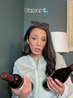 A woman with dark hair and sunglasses perched on her head holds two bottles of wine, looking directly at the camera with an open mouth as if speaking. Above her head is the text "Cancer" and a wave emoji. The background is a plain wall with a lamp in the background.