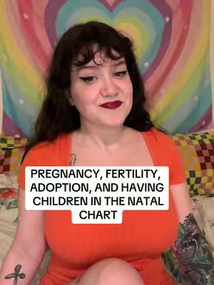 A woman with dark hair and red lipstick is centered in the frame, wearing an orange top. A text overlay in a white box reads, "PREGNANCY, FERTILITY, ADOPTION, AND HAVING CHILDREN IN THE NATAL CHART." The background is a tapestry with a rainbow design. The woman has tattoos on both arms. She is positioned indoors.