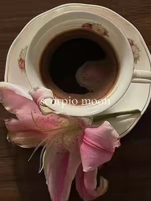 An overhead shot featuring a cup of coffee on a saucer, with a partially visible pink lily. The cup has dark coffee, and the saucer and cup feature floral designs. The lily is positioned near the cup, adding a soft pink contrast to the image. The words 'Scorpio moon' are overlaid over the cup.