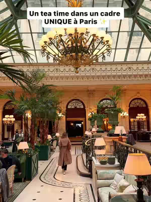 The image showcases the interior of an opulent building, likely a luxurious hotel, with a long hallway leading into the distance. The space is characterized by a grand chandelier, tall arched windows, and a high ceiling made of glass, suggesting a conservatory or atrium. A woman wearing a long coat is walking away from the camera.