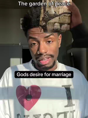A man with dark skin and short, styled hair is centered. He's wearing a white shirt with a large pink heart graphic and the letter "I." A book is held above his head, and a black rectangle with white text sits below the man's face.