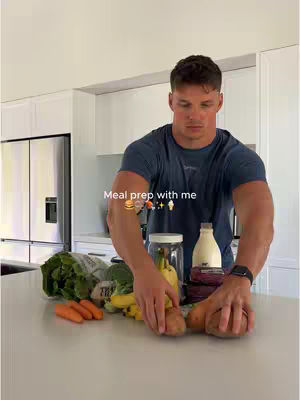 A man stands behind a counter with various fresh produce and ingredients, appearing to prepare a meal. He is wearing a dark blue t-shirt and has short brown hair. The background is a modern kitchen with white cabinets and a stainless steel refrigerator. Text overlayed on the image reads 'Meal prep with me' along with food-related icons.