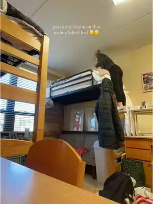 A college dorm room with a lofted bed is shown. A person is standing on the bed, looking at the top bunk, which has text overlaid on it. The room features a window, a desk, and some additional furniture and items.