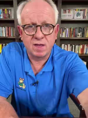 A man wearing glasses and a blue polo shirt speaks directly to the camera. He is in front of a bookshelf.