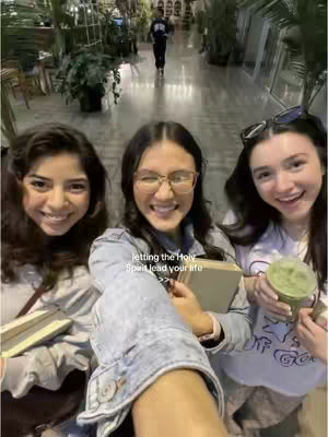 A selfie-style shot of three young women smiling and looking at the camera. The central figure is holding the camera, with the other two on either side. The background shows an indoor setting with lots of greenery. In the background, someone is walking towards the camera.