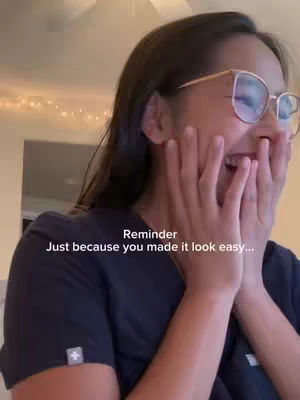 A young woman with long dark hair and glasses is shown from the chest up. Her hands are clasped over her mouth, eyes widened, looking surprised or emotional. The background is soft and blurred, with warm lighting.