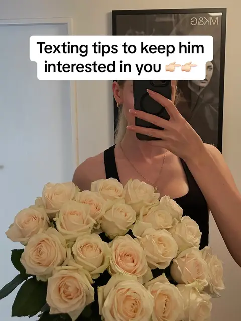 A woman holds a large bouquet of roses while taking a mirror selfie. The background features a wall, door, and a piece of framed art. A text overlay at the top reads 'Texting tips to keep him interested in you' with two pointing finger emojis.