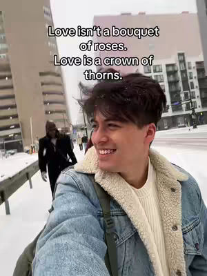 A young man smiles broadly in a selfie-style shot, with a snowy urban street scene in the background. He wears a light-colored sweater under a denim jacket with a tan-colored fleece lining around the collar. The background features several tall buildings and a person wearing a black coat walking away.
