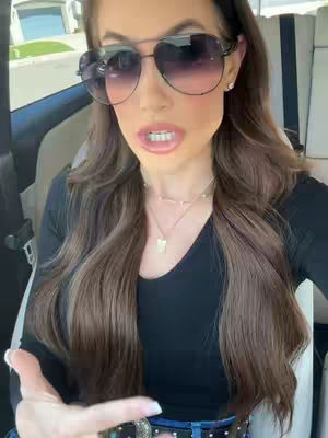 A woman with long, wavy brown hair and large sunglasses is sitting in a car. She is looking directly at the camera with a slightly open mouth. The background shows the car's interior and some glimpses of the outside environment.